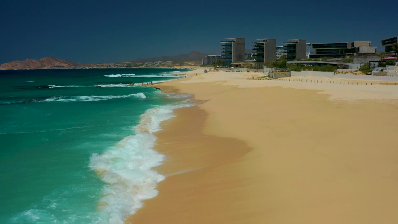 A diagonal surf line runs along a wide beach with turquoise water offshore.