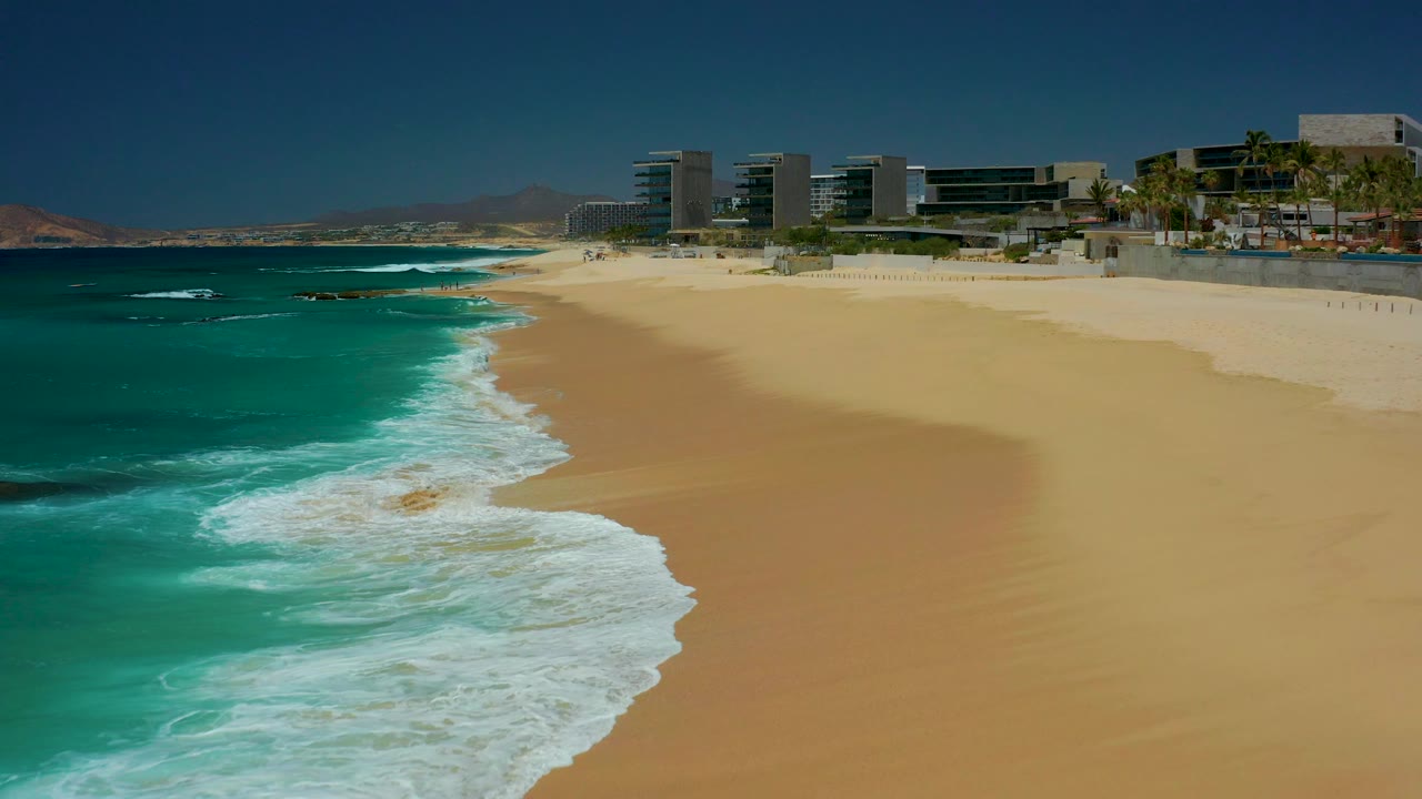 Opening frame of the Dreams Los Cabos beach, with turquoise surf at left, golden sand across the foreground, modern beachfront buildings in the distance, and no visible people.