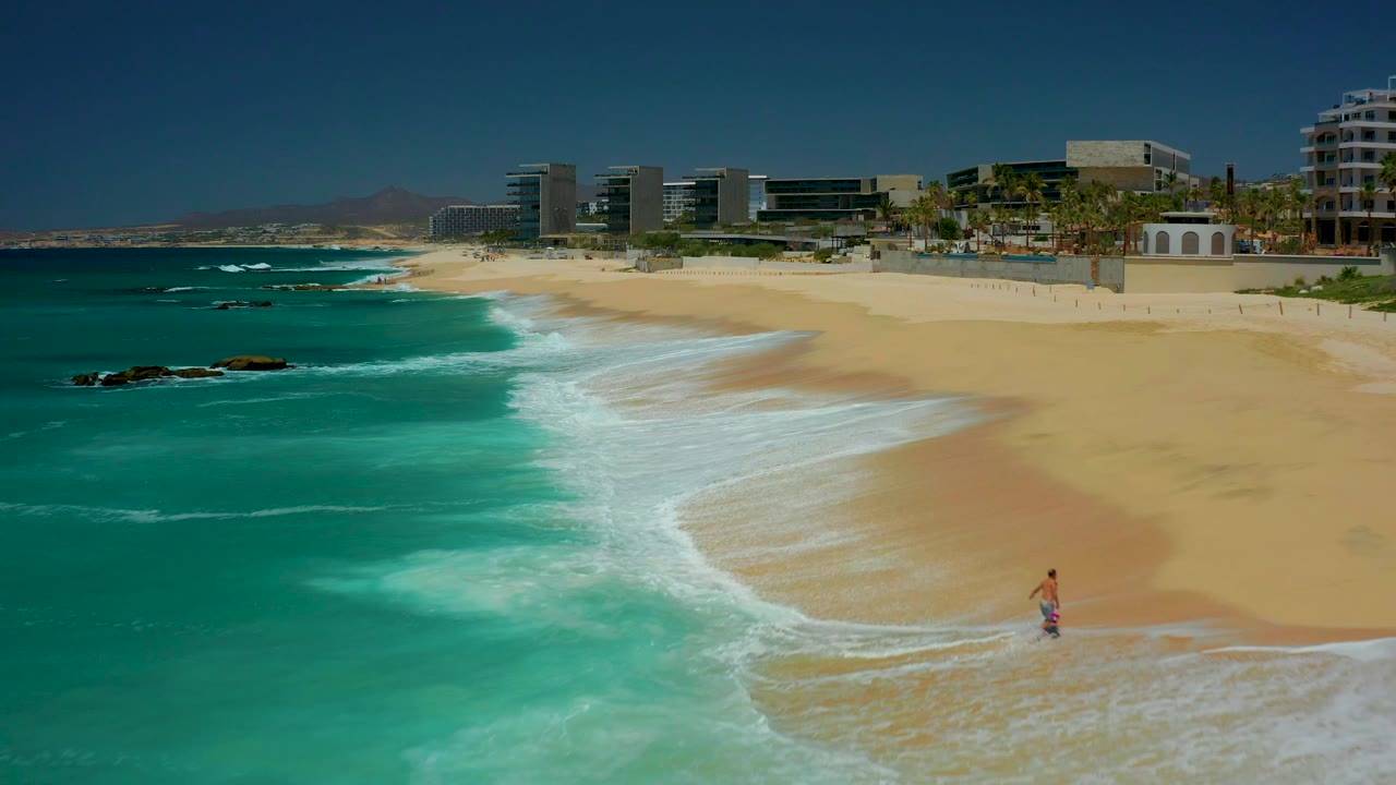 Wide aerial view at Dreams Los Cabos, Mexico, showing turquoise ocean washing onto broad sand, beachfront buildings and palm trees in the distance, and a small man standing or walking near the lower shoreline.