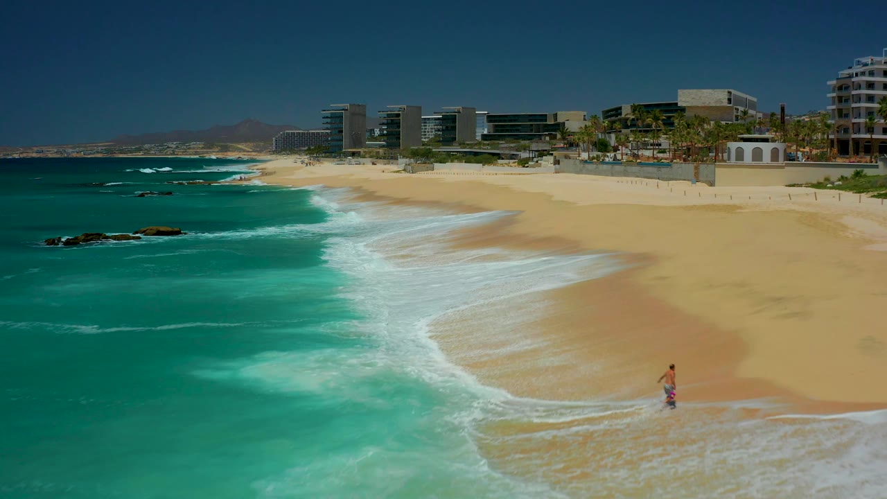 Aerial view at Dreams Los Cabos, Mexico, with the surf line curving across the sand, resort buildings and palms along the back of the beach, and a small man farther right near the waterline.