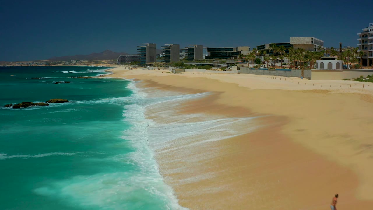 Dreams Los Cabos shoreline view with bright sand, turquoise water, white foam bands, distant beachfront buildings and palms, and the small man partly cut off at the lower right edge.