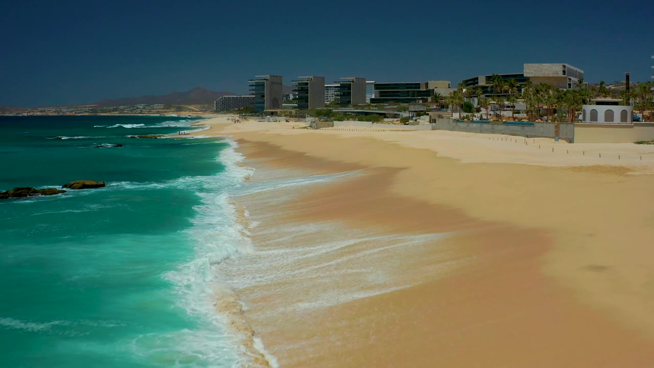 Empty sandy beach aerial with turquoise surf, rocks offshore, resort buildings, palms, and clear sky.