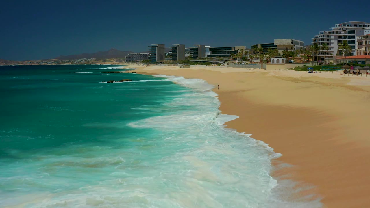 White surf spreads farther along the diagonal sandy shoreline below beachfront buildings.