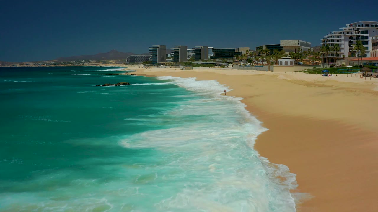 At 0:02, a curved sheet of white foam pushes up the Dreams Los Cabos beach while modern beachfront buildings, palms, and open blue sky line the upper frame.