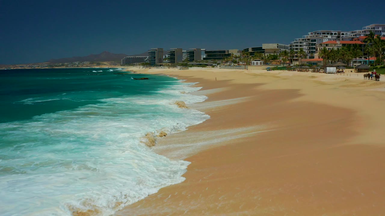 At Dreams Los Cabos in Baja California Sur, turquoise water breaks onto tan sand with resort buildings and tiny beachgoers visible near the upper right.