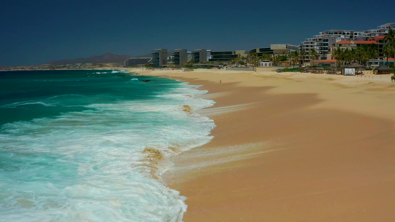 White water fills more of the lower left foreground at Dreams Los Cabos, with broad empty sand, resort buildings, and a few tiny distant figures near the upper right.