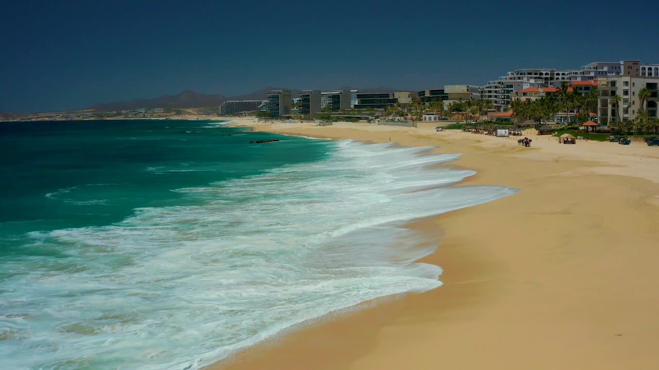 A second view of Dreams Los Cabos shows a broad band of white surf curling across golden sand, with beachfront buildings and palms along the upper right, with no visible people.