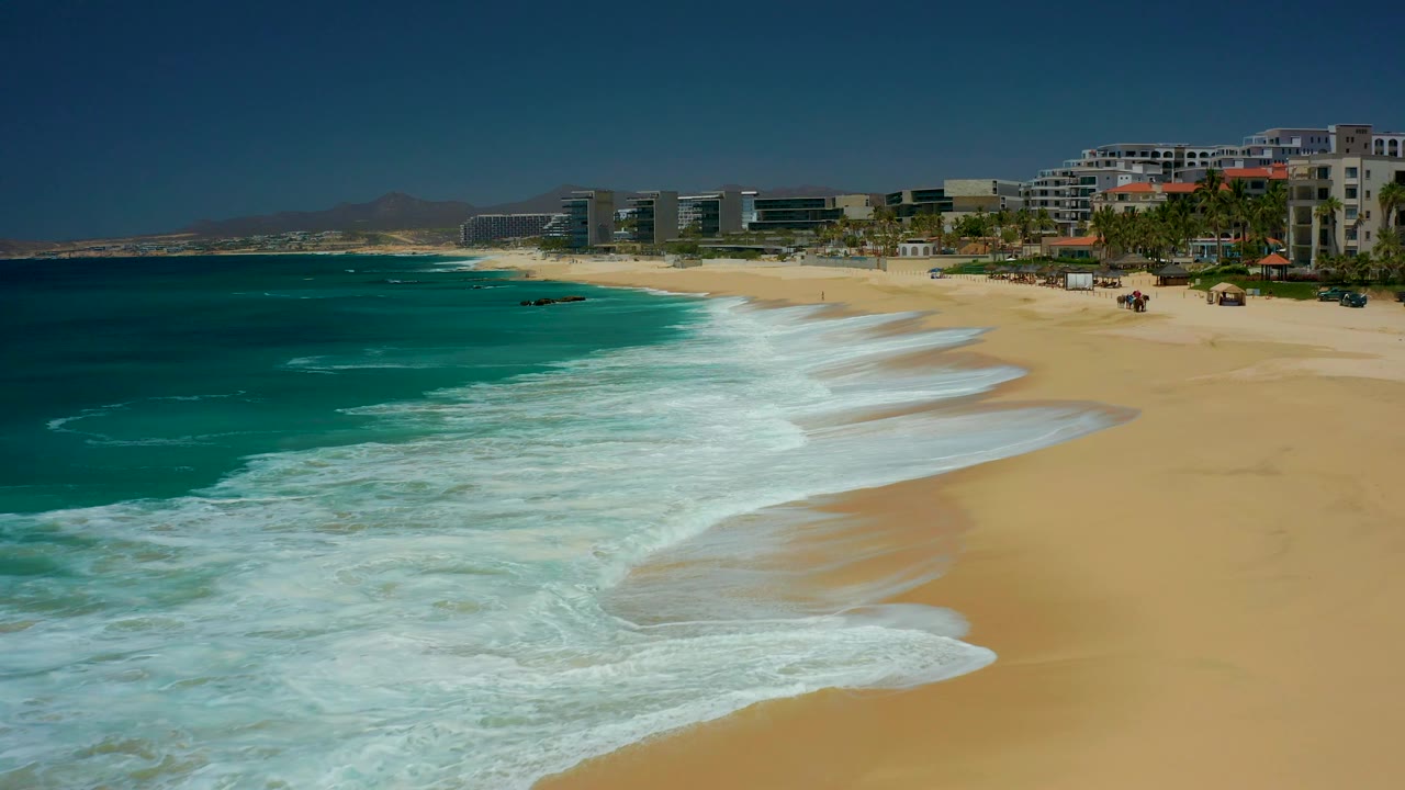 At Dreams Los Cabos, the foam sheet reaches farther up the shoreline while the turquoise water darkens offshore and the resort buildings remain in the background, with no visible people.