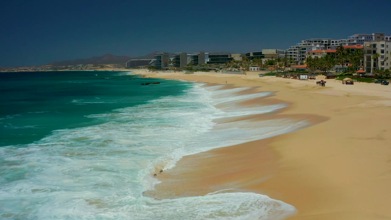 The Dreams Los Cabos shoreline appears wider as foamy water recedes over wet sand, with blue-green ocean on the left and resort structures along the upper right, with no visible people.