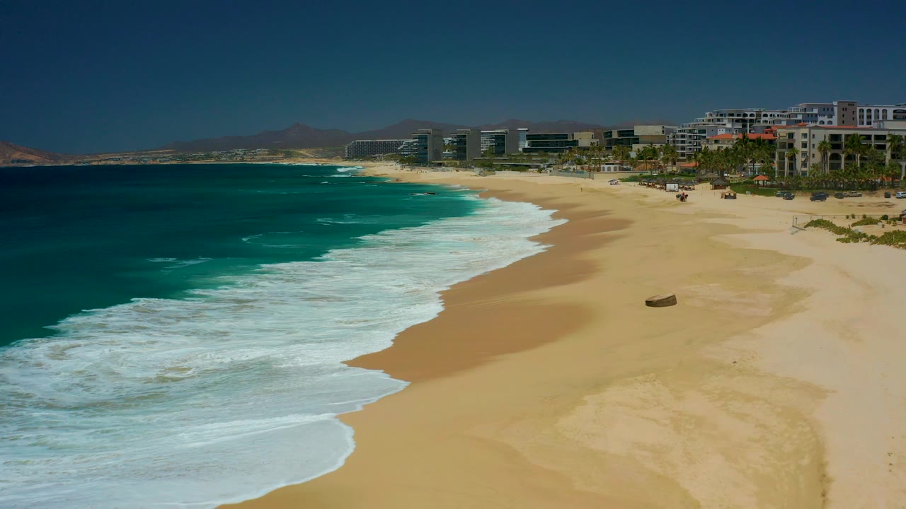 Turquoise ocean waves reaching a sandy beach with distant coastal buildings.