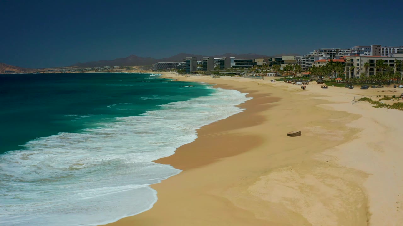 White foam spreads farther across the Dreams Los Cabos shoreline, with turquoise water on the left, tan sand on the right, distant buildings, and no visible people.