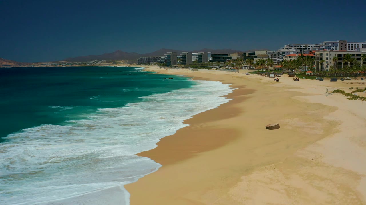 The surf forms a curved white band along the sandy beach at Dreams Los Cabos, with a small round object on the sand, distant buildings, and no visible people.