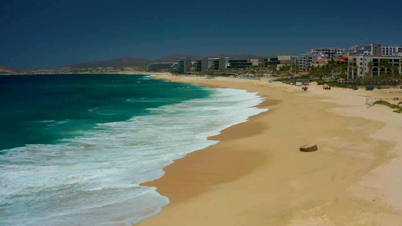 An elevated view shows the turquoise ocean and white wash moving along the Dreams Los Cabos beach, with the small beach object lower on the sand and no visible people.