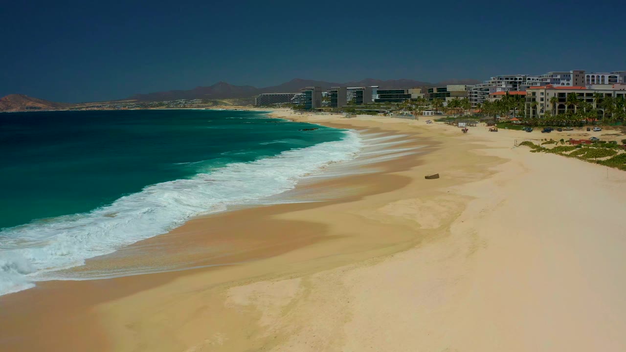 A slightly later view of the Dreams Los Cabos shoreline shows a wider band of white foam reaching up the beach, with tan sand, beachfront buildings, and distant mountains, no visible people.