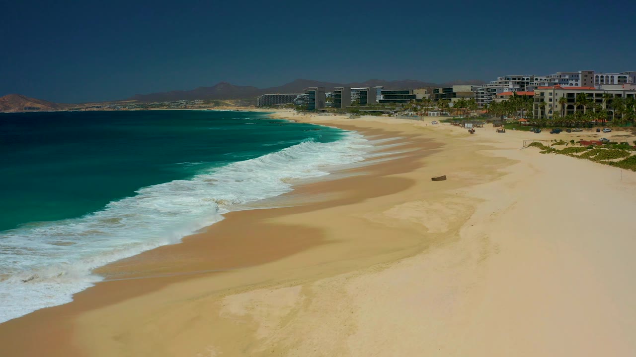 A broad section of Dreams Los Cabos beach fills the frame as white waves approach the sand, with a small dark object on the beach, resort buildings, palms, and mountains in the background, no visible people.
