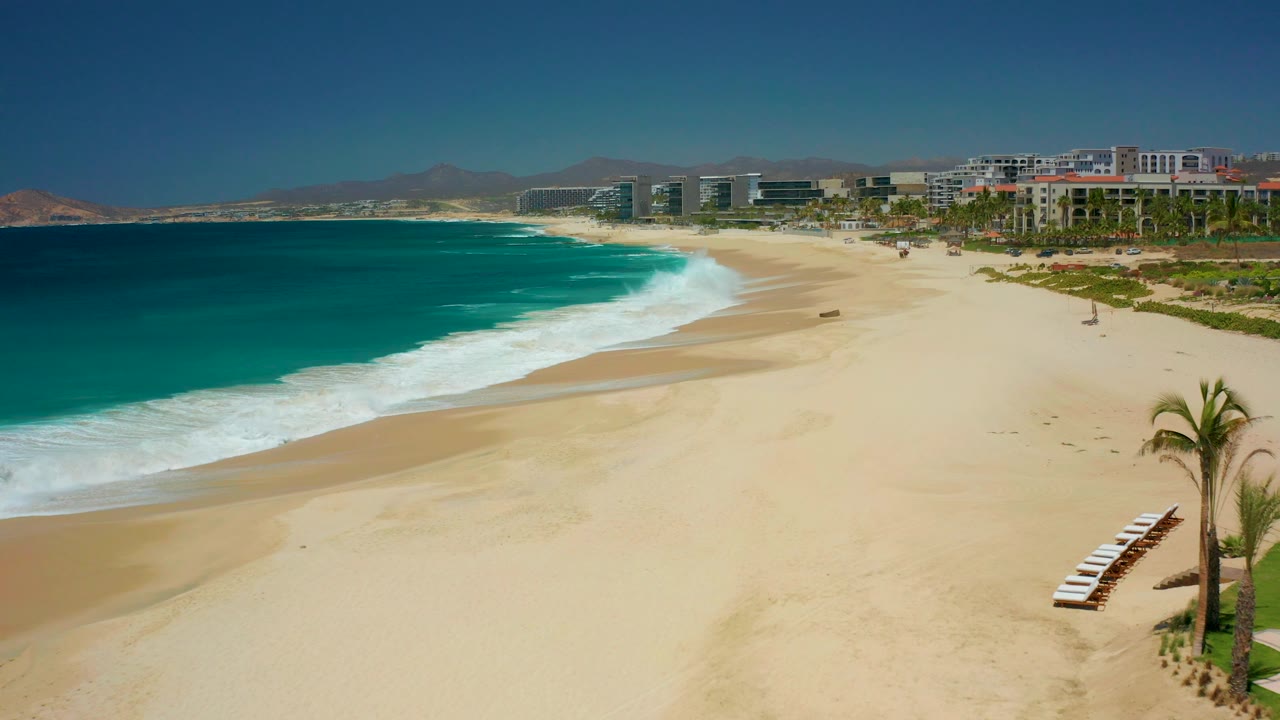 At Dreams Los Cabos in Baja California Sur, turquoise water approaches a broad sandy beach with lounge chairs and a palm tree at lower right, resort buildings in the background, and no visible people.