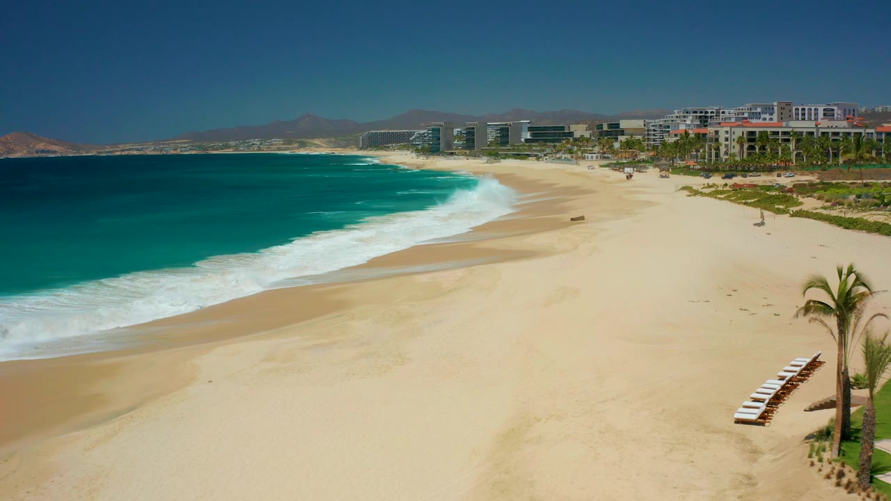 At Dreams Los Cabos in Baja California Sur, a curved band of white surf spreads across the sand while lounge chairs and a palm tree sit near the lower right edge and no visible people appear.