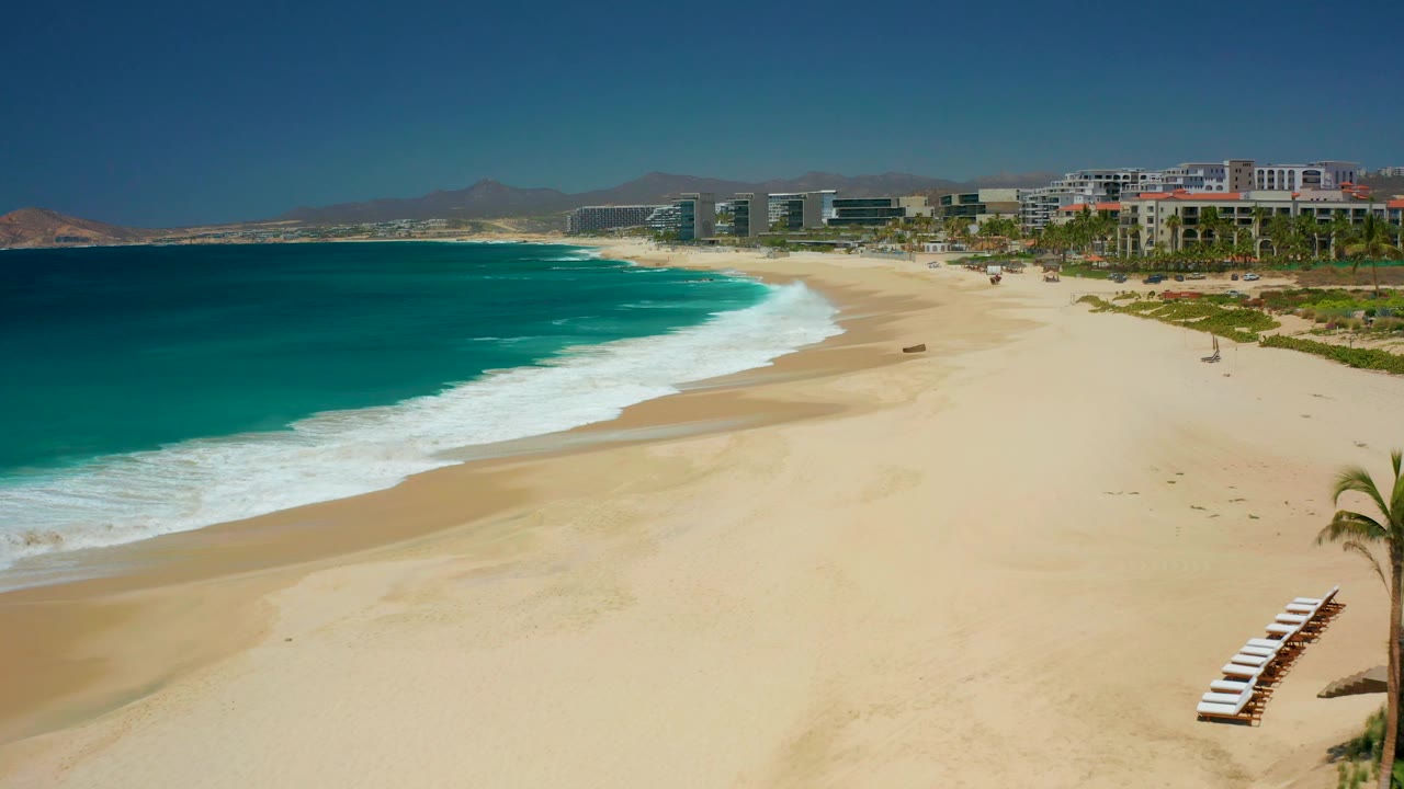 At Dreams Los Cabos in Baja California Sur, the camera view shows more open sand to the right, turquoise ocean to the left, a white foam line near shore, distant buildings, and no visible people.