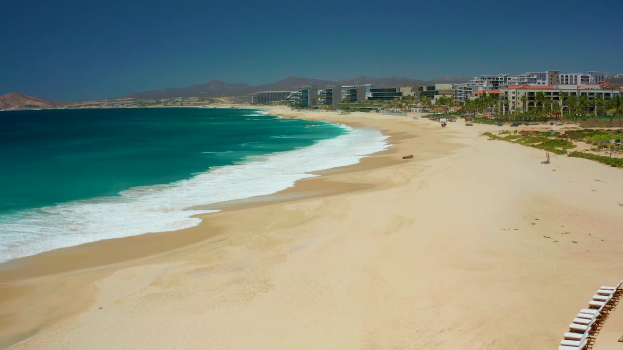 Turquoise water and white surf border a broad sandy beach with buildings beyond.