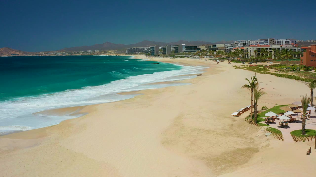 At Dreams Los Cabos in Baja California Sur, turquoise surf approaches a wide sandy beach with palms and sun umbrellas at right, resort buildings beyond, and no visible people.