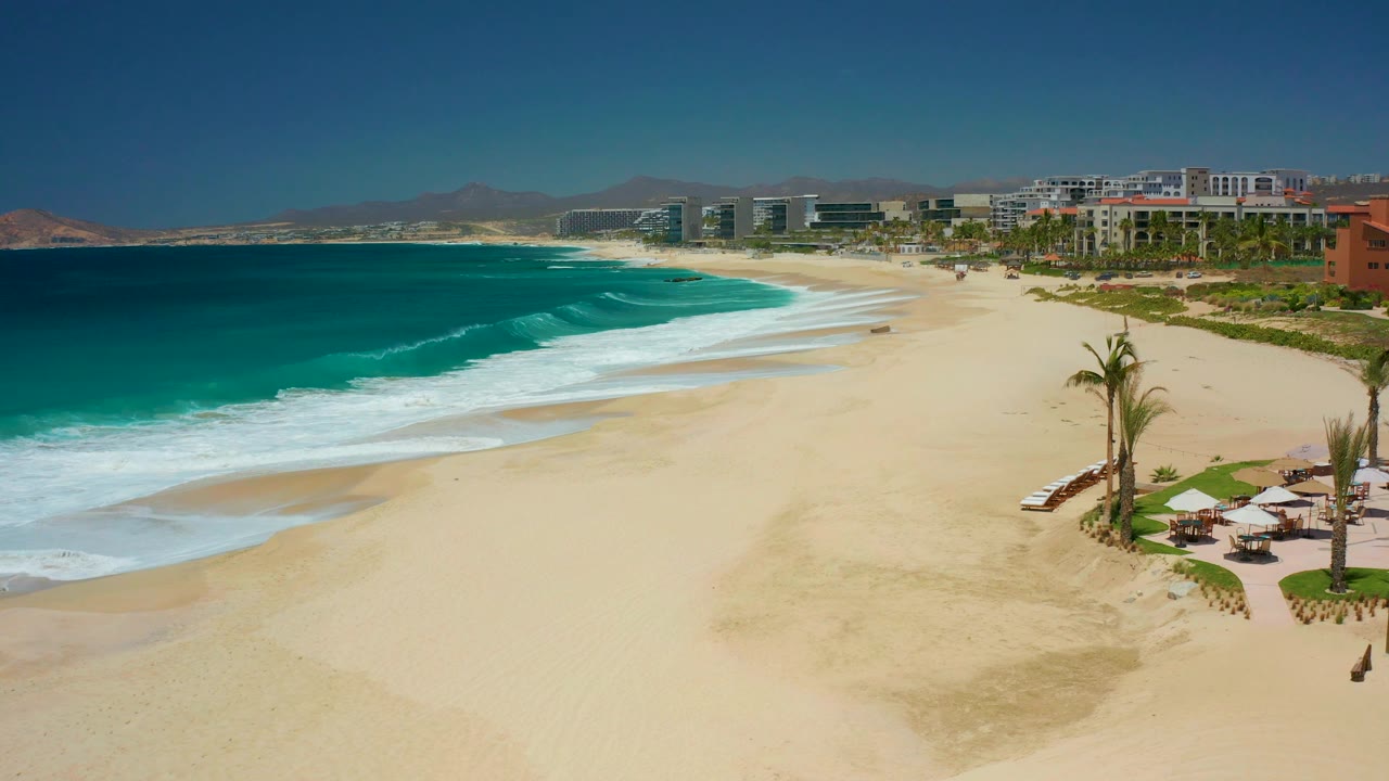 At Dreams Los Cabos in Baja California Sur, white foam spreads along the shoreline below a clear sky, with palms, umbrellas, resort buildings, distant mountains, and no visible people.