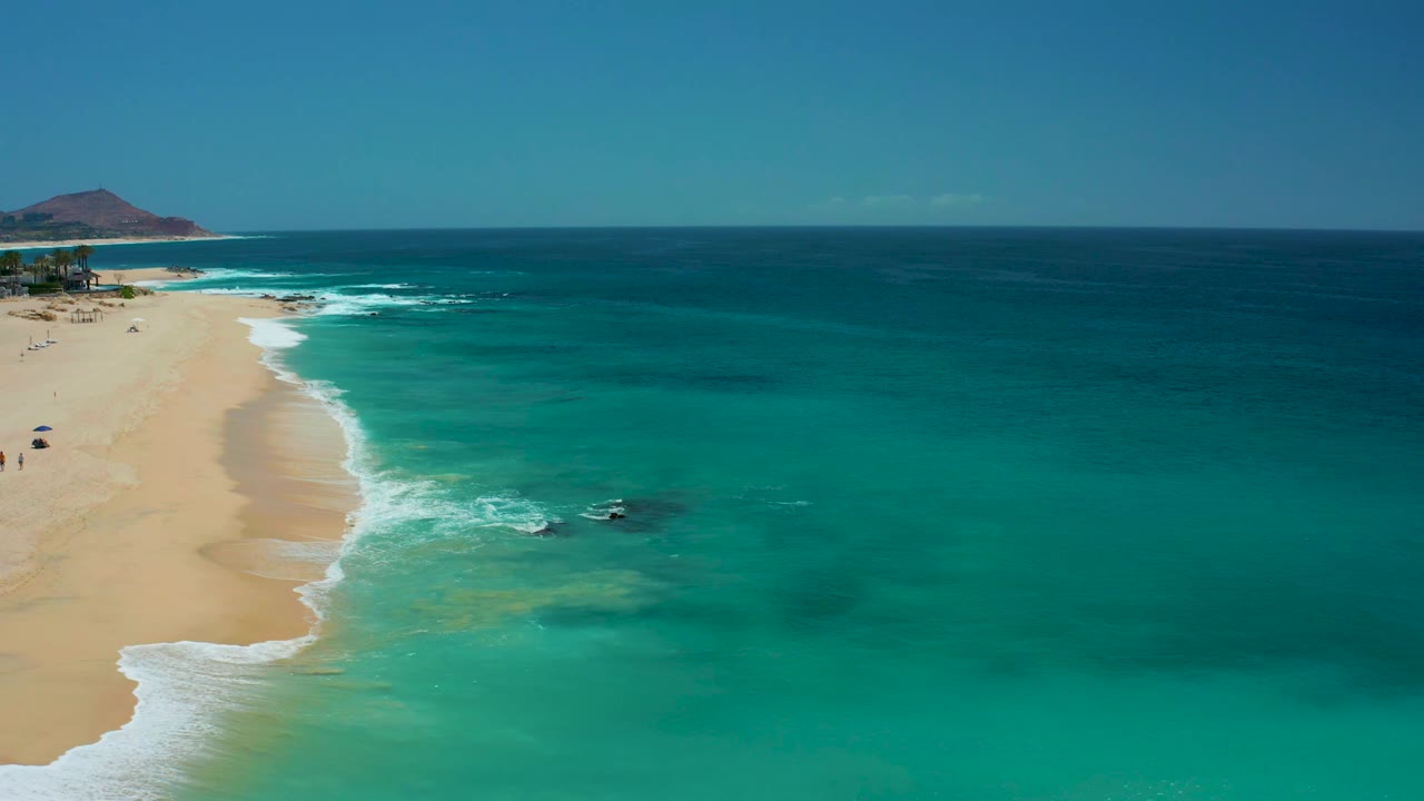 Aerial frame from Dreams Los Cabos with the shoreline curving along the left side, white foam washing onto sand, turquoise shallows in the center, darker ocean beyond, and no visible people.