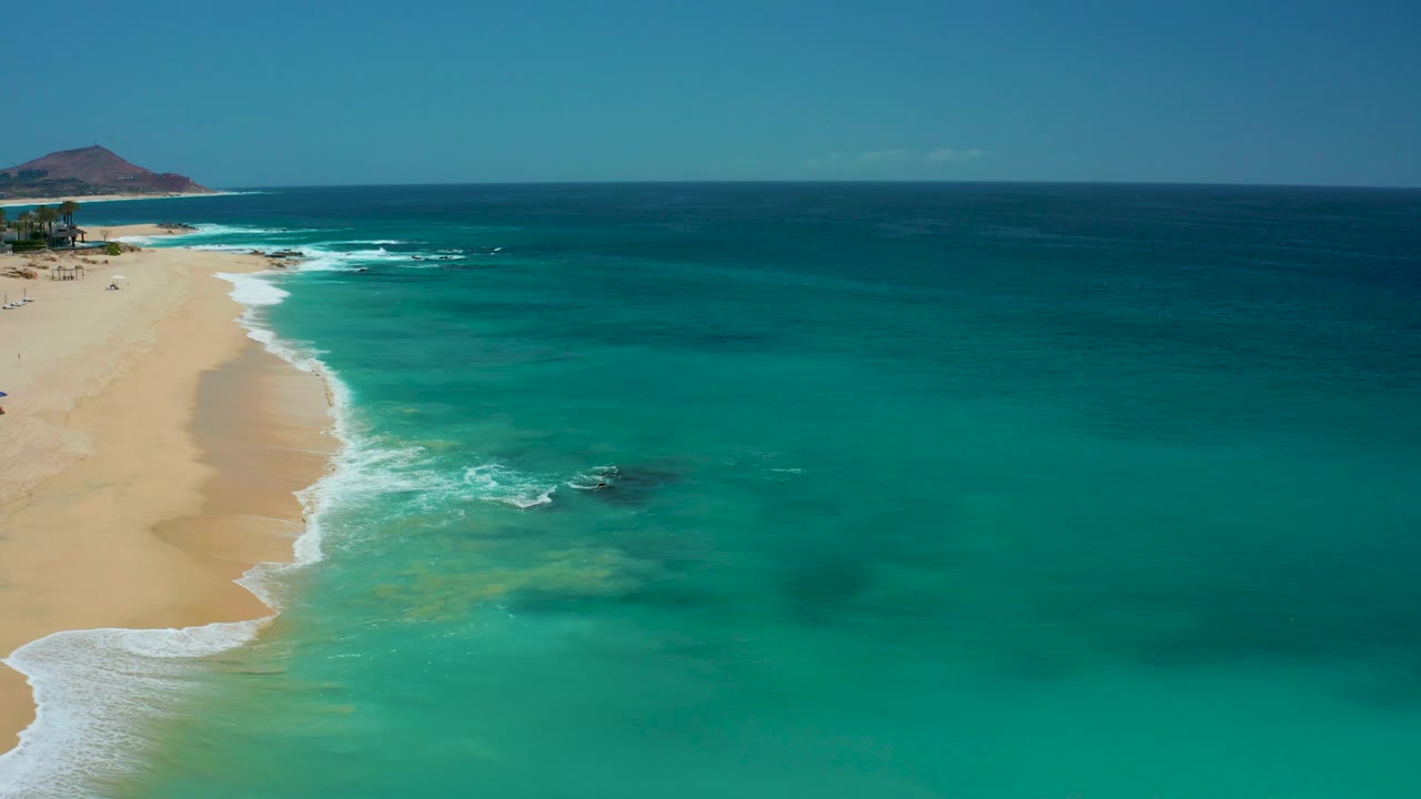 A Dreams Los Cabos aerial frame with a narrow strip of sand on the left, broad turquoise ocean filling the center and right, scattered white wave breaks near shore, and no visible people.
