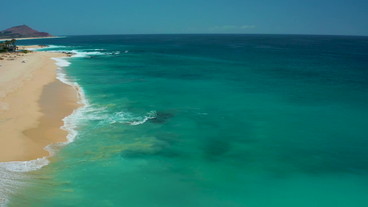Aerial view over Dreams Los Cabos with sandy beach along the left border, a bright foam line in the lower left, shallow green-blue water near shore, darker sea near the horizon, and no visible people.