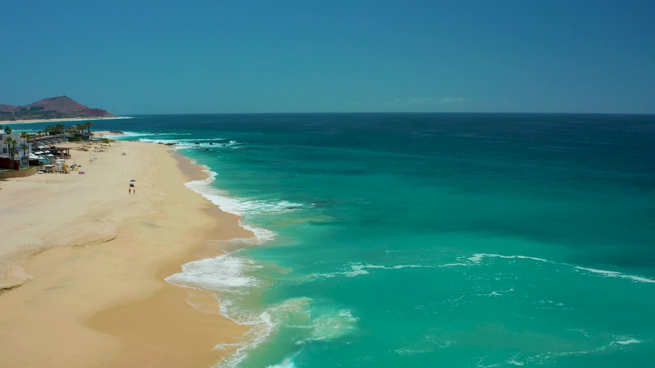 Opening aerial view at Dreams Los Cabos showing pale sand on the left, turquoise ocean on the right, white surf along the shoreline, distant beach structures, and tiny walkers near the water.