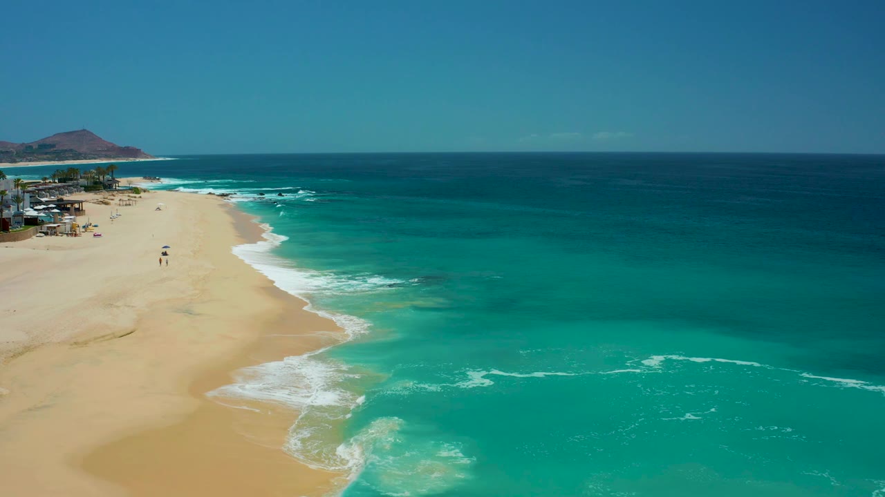 Aerial view at Dreams Los Cabos where the beach occupies more of the left frame, the ocean remains bright turquoise, and two small walkers appear near the mid-beach surf line.