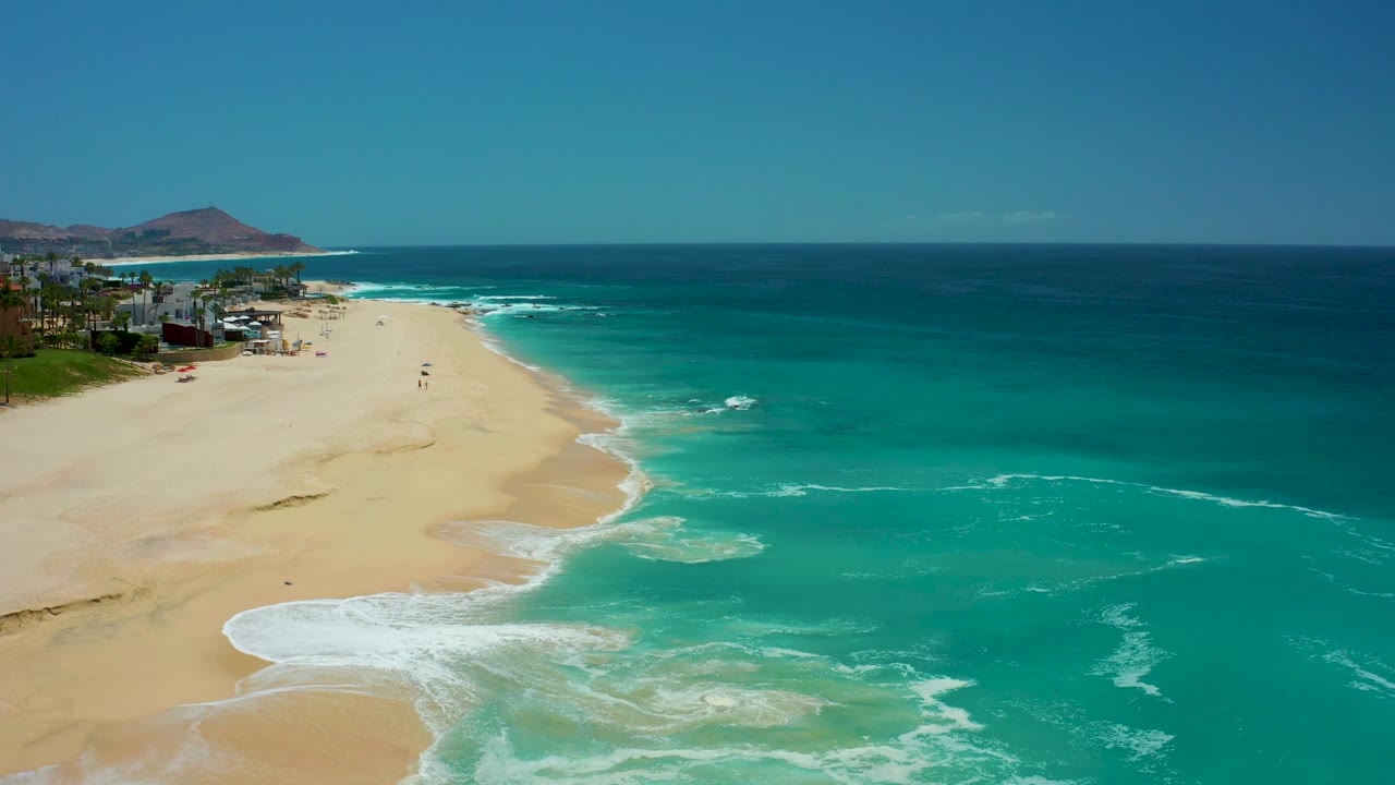 Aerial view over Dreams Los Cabos with a pale sand beach on the left, turquoise shallows and white foam in the foreground, darker blue sea beyond, distant hills, and no visible people.