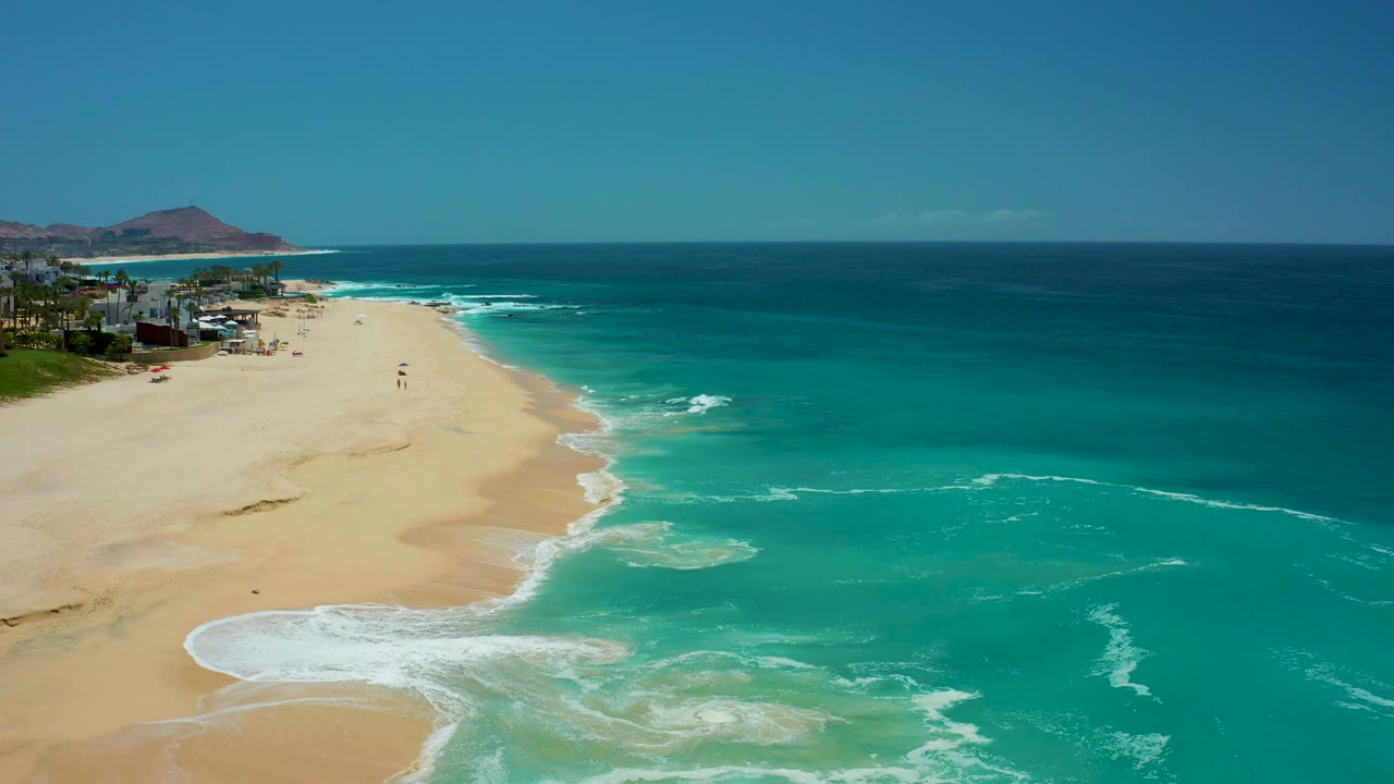 At Dreams Los Cabos, a curving surf line meets smooth sand while the beach settlement stays at the far left edge, with open sea filling the right side and no visible people.