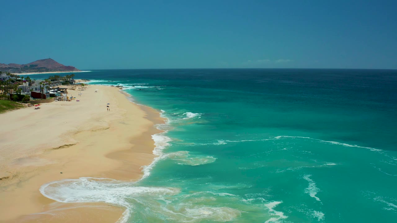 A high coastal view at Dreams Los Cabos shows a broad wedge of sand, thin white foam bands, turquoise water across the lower right, distant hills, and no visible people.