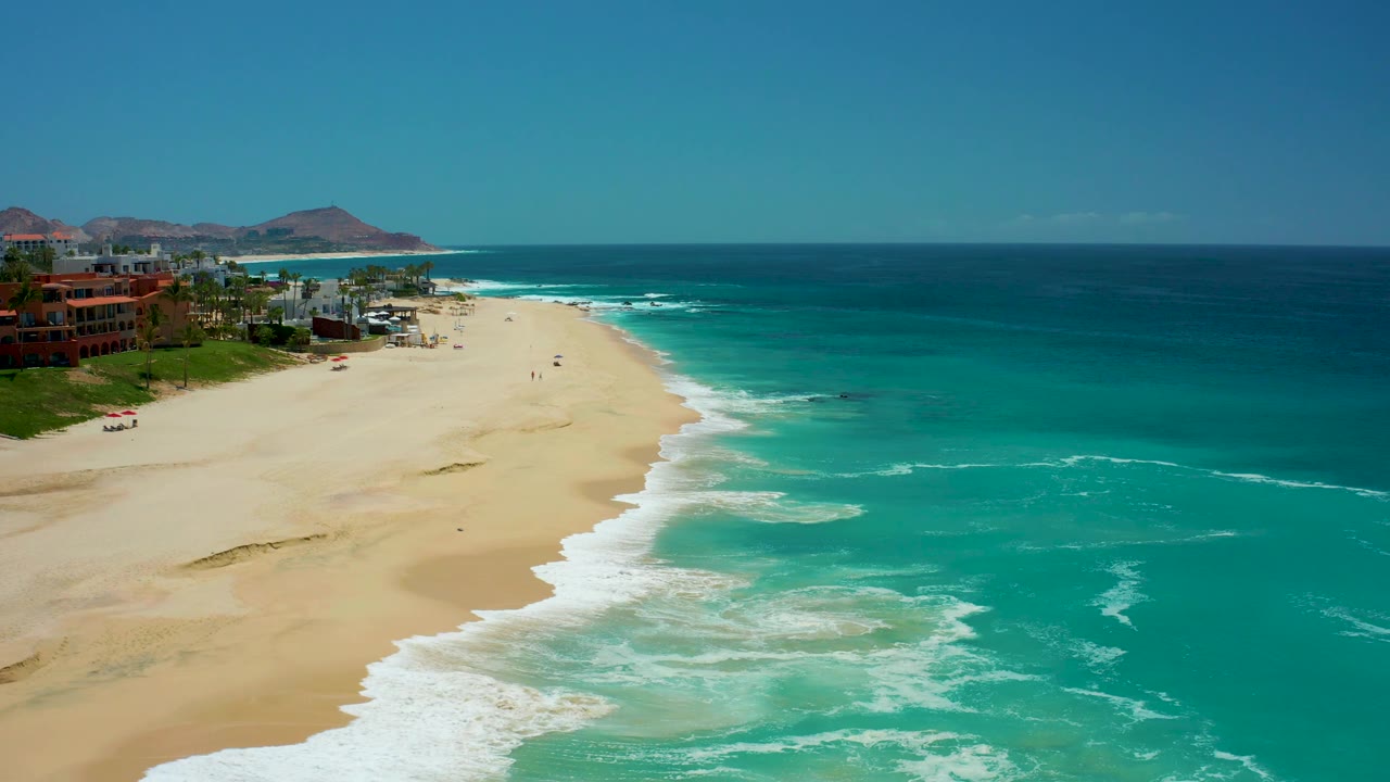 At Dreams Los Cabos in Baja California Sur, a curling band of white surf traces the beach while resort structures remain along the left edge under clear sky, no visible people.