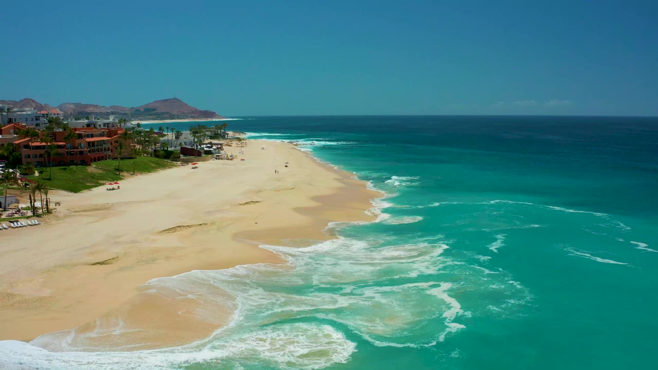 At 0:01, the Dreams Los Cabos shoreline shows a broad sandbar, turquoise water to the right, foam patterns advancing near the beach, and no visible people.