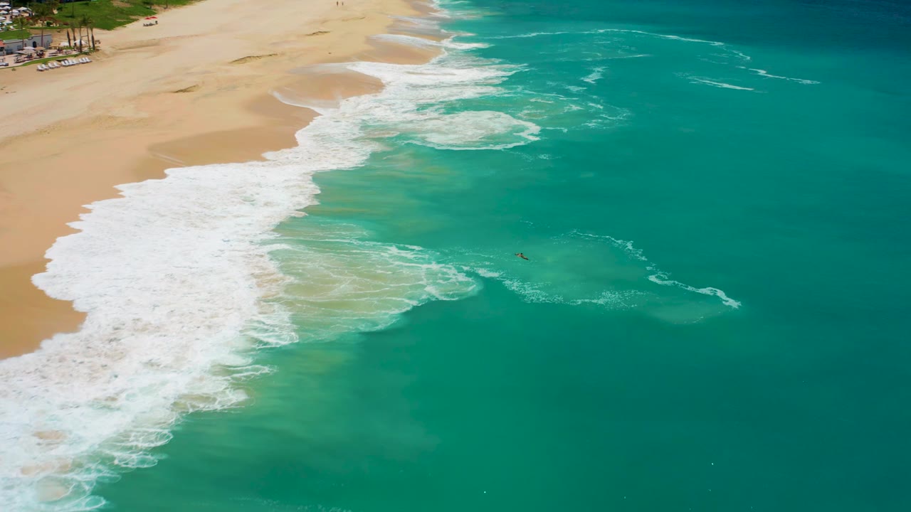 Overhead view at Dreams Los Cabos of white waves spreading across tan sand with turquoise water filling the right side and a tiny distant swimmer in the shallows.