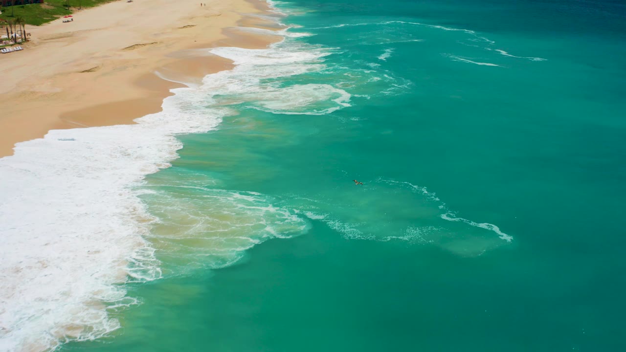 Overhead Dreams Los Cabos frame where white surf fans out from the left edge, clear turquoise water covers most of the image, and a small swimmer is visible near the right side.