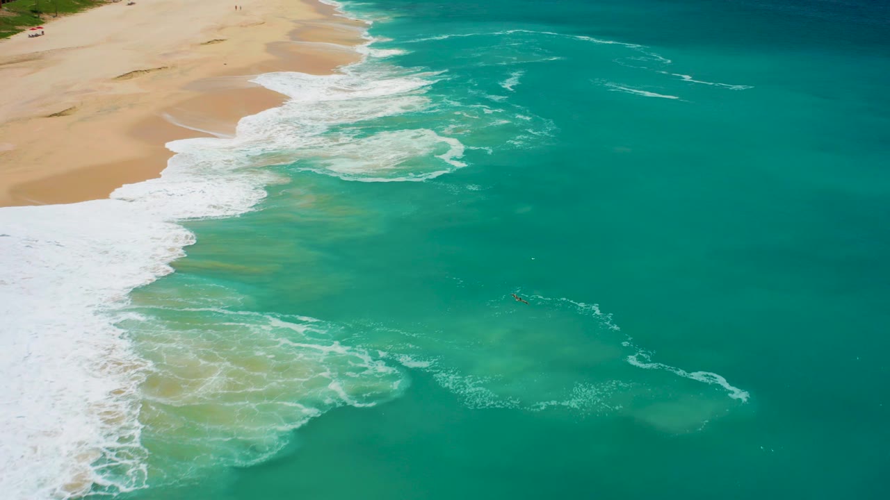 Foamy waves break along a sandy shoreline seen from above.