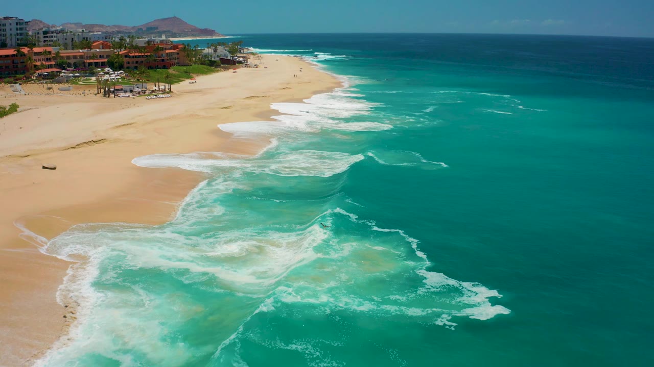 Aerial frame at Dreams Los Cabos, Mexico, showing foamy wave lines advancing toward pale sand with beachfront buildings and palms along the upper left, no visible people.