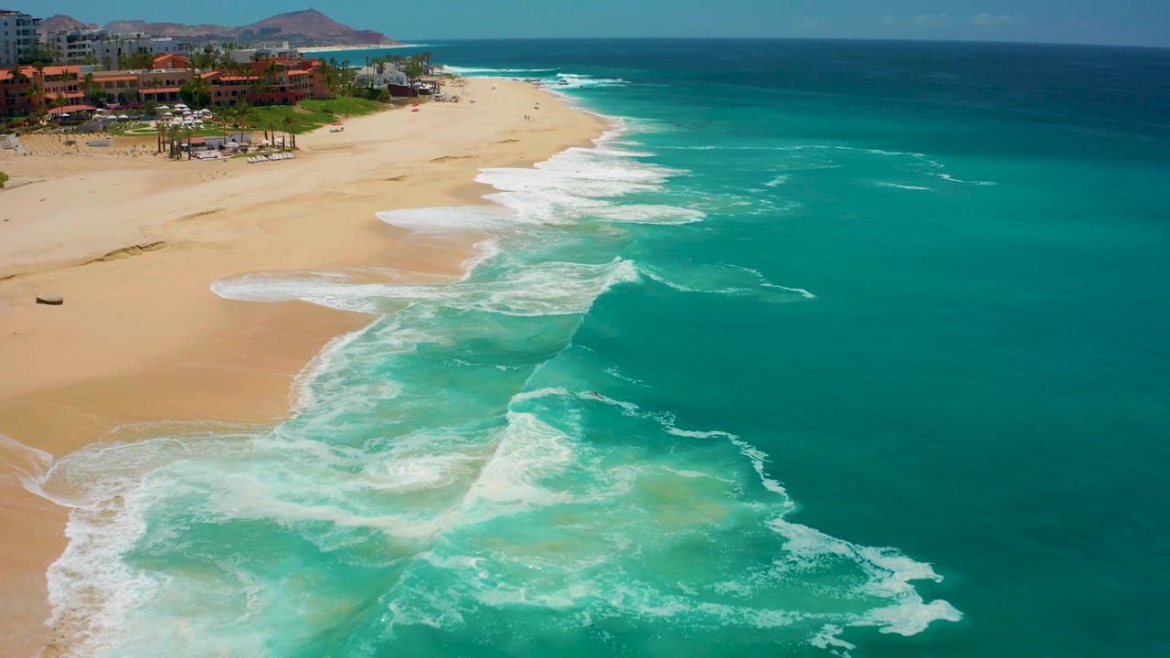 Aerial view at Dreams Los Cabos, Mexico, with more open turquoise water filling the right side and white foam breaking along the sandy beach to the left, no visible people.