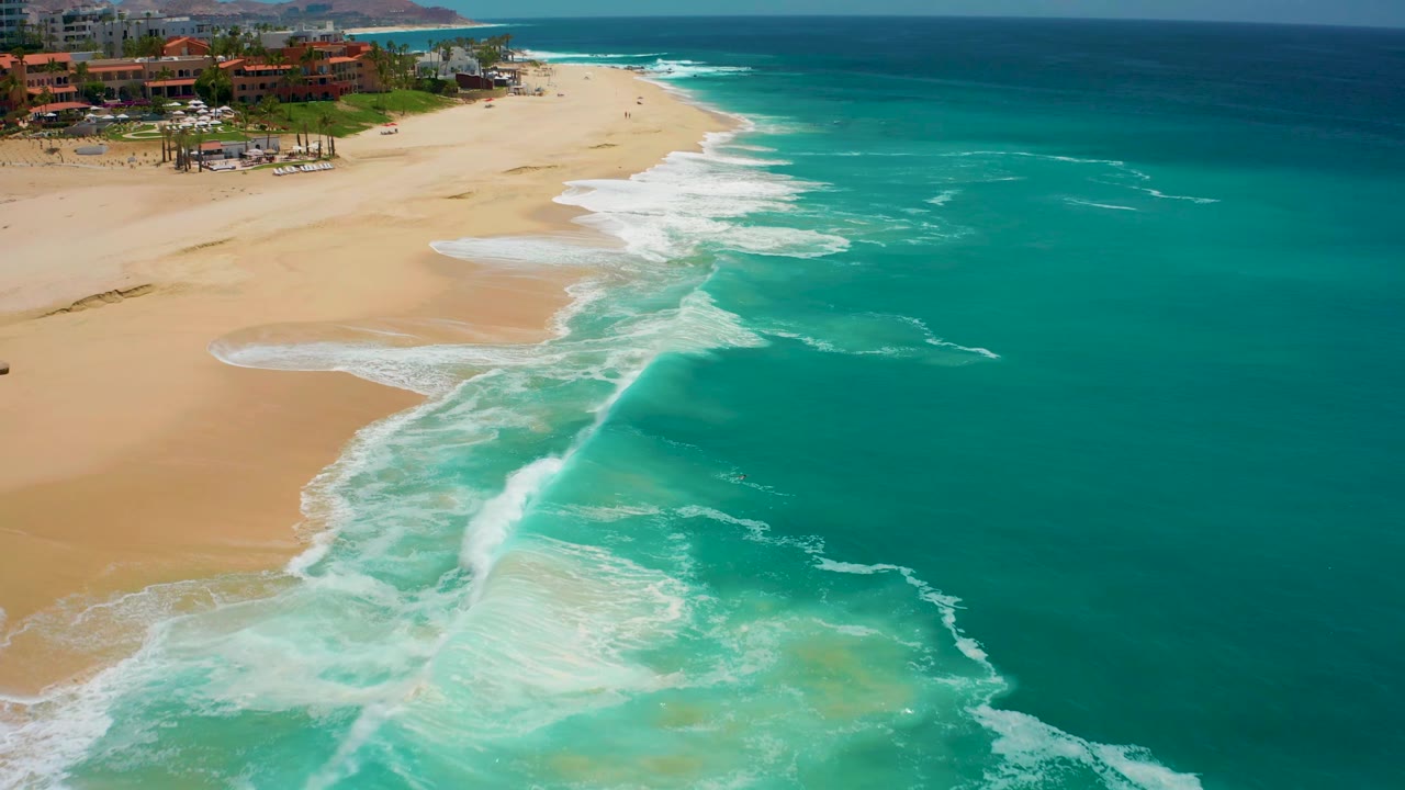 Aerial frame at Dreams Los Cabos, Mexico, showing a diagonal band of white surf crossing shallow turquoise water beside a wide sandy beach, no visible people.
