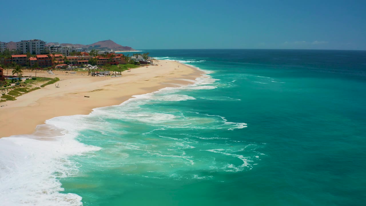 At Dreams Los Cabos, a bright foamy wash reaches the lower left shoreline while turquoise water fills the frame and the resort edge remains in the left background with no visible people.