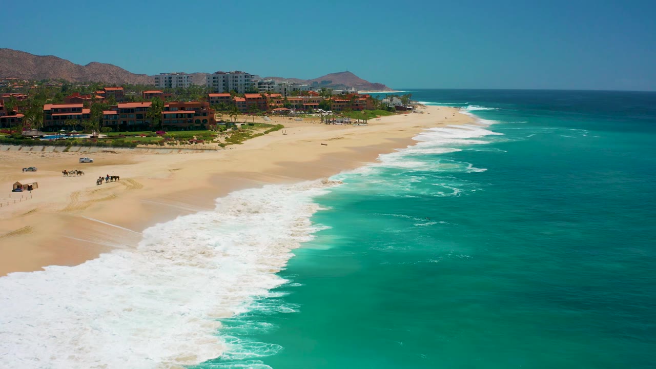 White surf spreads across a sandy resort beach with turquoise water offshore.