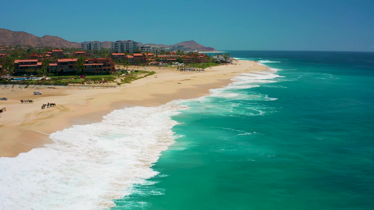 At Dreams Los Cabos, the beach curves toward the distance with resort buildings on the left, turquoise sea on the right, white surf bands, mountains behind the buildings, and small people on the sand.