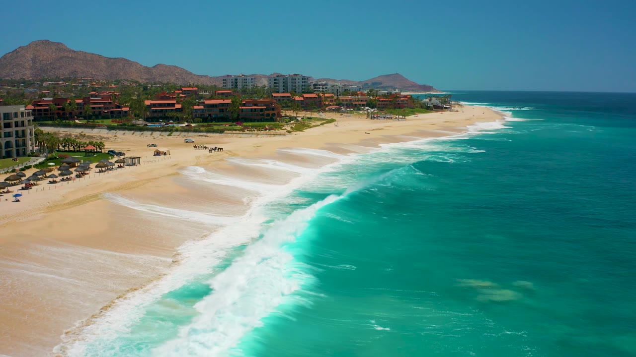 Aerial frame at Dreams Los Cabos with the surf pushed farther across the lower shoreline, resort structures along the upper beach, and clear blue sky above dry hills.