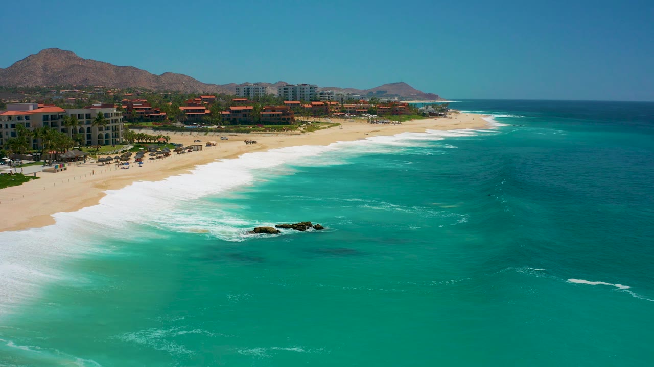 Opening aerial frame over Dreams Los Cabos with turquoise water in the foreground, white surf reaching the sand, coastal buildings along the beach, mountains behind them, and no visible people.