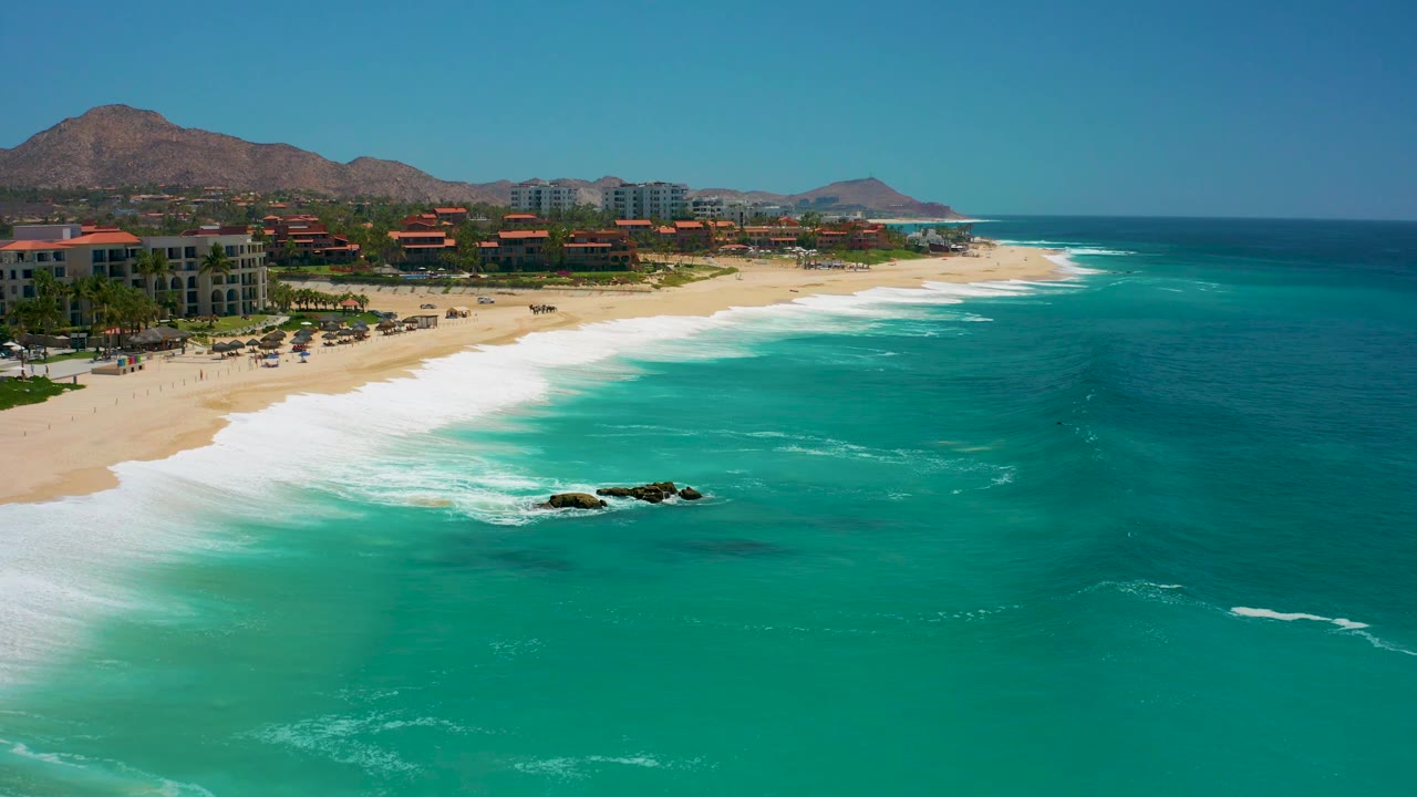 Wide aerial coastline with white waves breaking on sand and resort buildings below mountains.