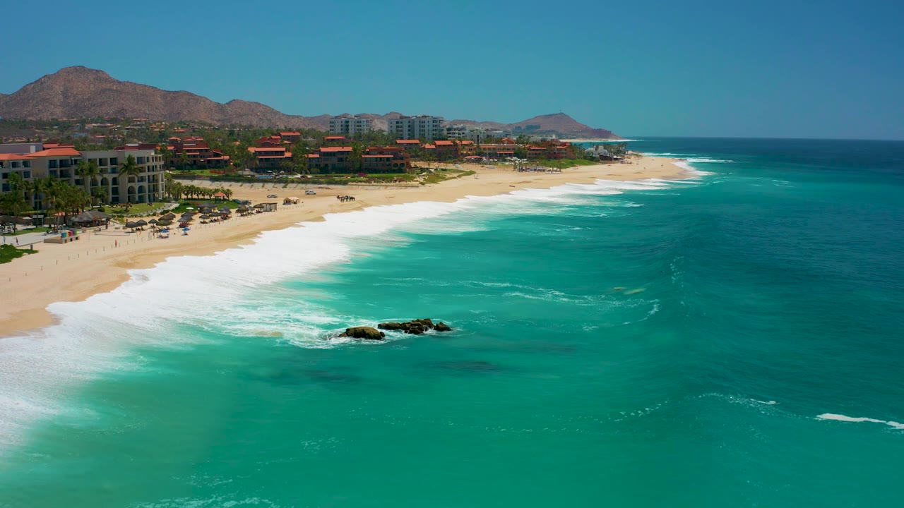 The frame shows Dreams Los Cabos from above with more open turquoise water to the right, white surf curling along the beach, coastal buildings set back from the sand, mountains beyond, and no visible people.
