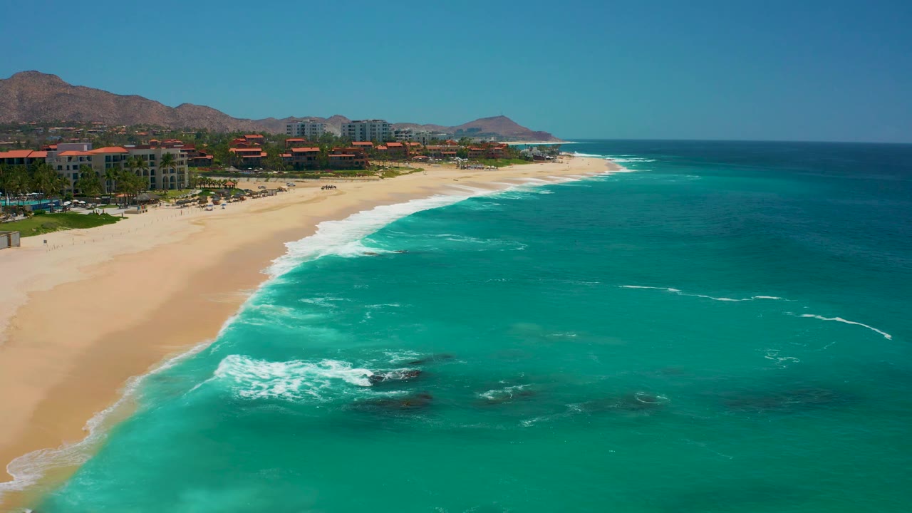 At Dreams Los Cabos, white foam advances over the pale sand while the resort buildings remain along the left side, with mountains under a clear sky and no visible people.