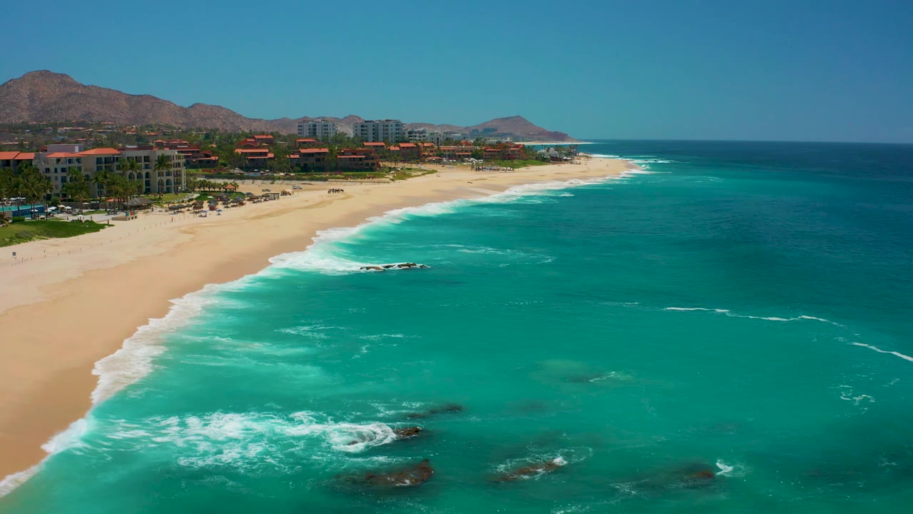 Aerial view at Dreams Los Cabos shows white water curling around dark rocks near shore, with a sandy beach, resort buildings, mountains, and no visible people.